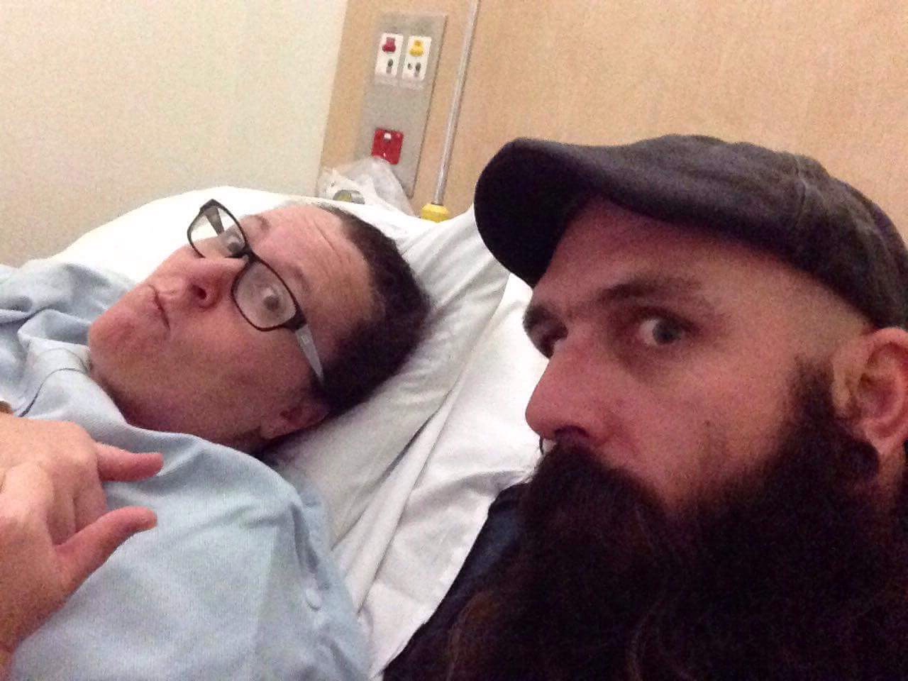 A woman on a bed in a hospital gown gives a thumbs up with a bearded man in a dark hat