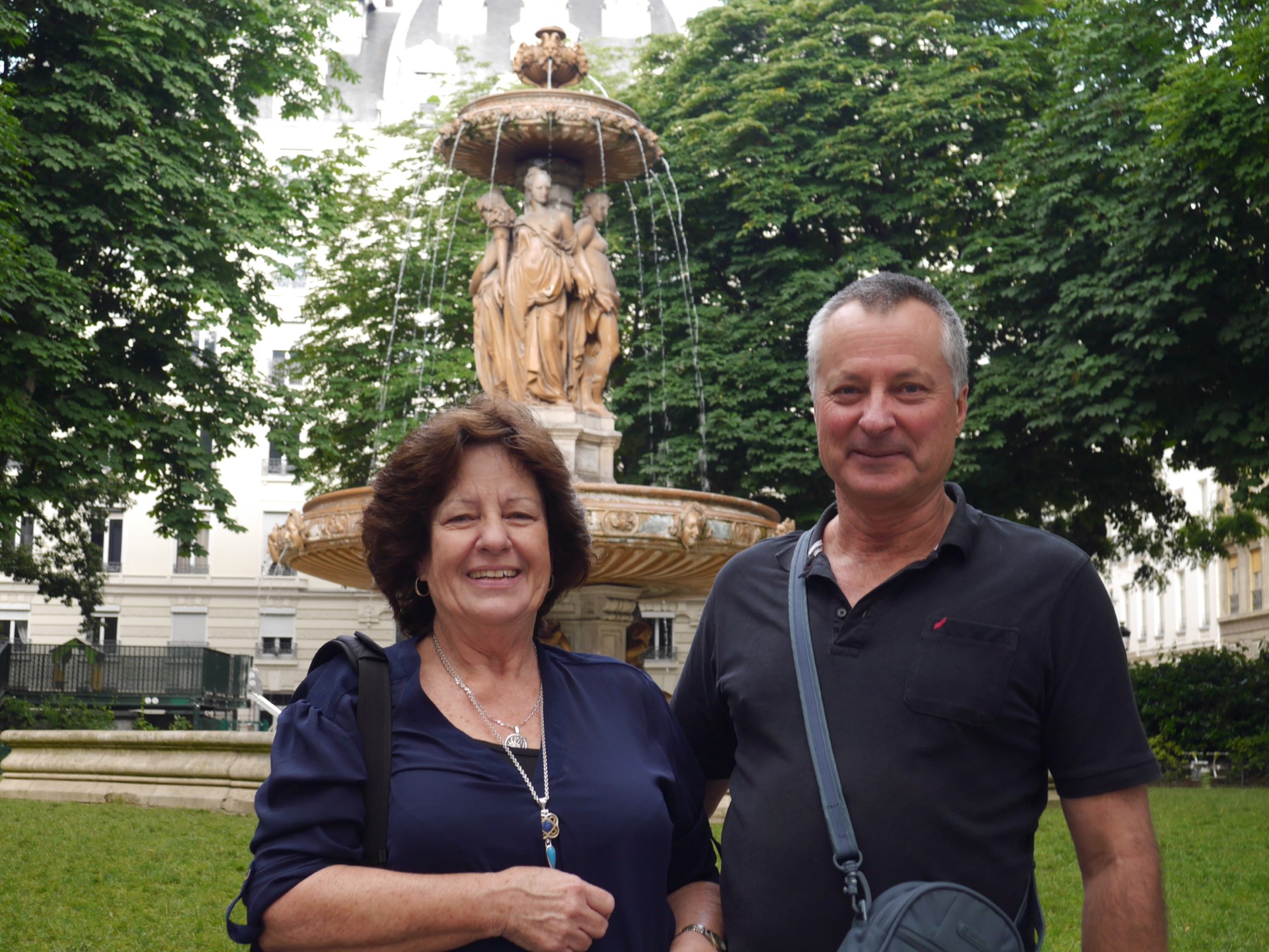 A man stands with his arm around a woman in a park in front of a fountain. They're both smiling.