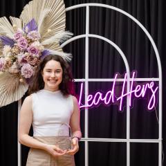 Dr Clare Mahon standing in front of a LeadHers sign holding her award. 